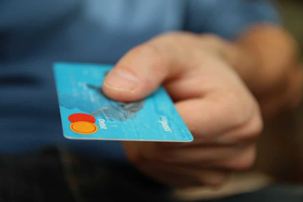 O persoană ține un card de credit în mâna dreaptă. Cardul are un fundal albastru cu un cerc portocaliu și roșu pe el. Unghia persoanei poate fi văzută în prim-plan. Degetul lor arătător se sprijină pe partea de sus a cardului, în timp ce degetul mare îl ține de dedesubt.